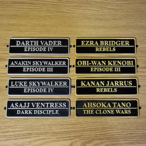 May include: Six black and gold rectangular nameplates with gold lettering. The nameplates feature characters from the Star Wars franchise, including Darth Vader, Ezra Bridger, Anakin Skywalker, Obi-Wan Kenobi, Luke Skywalker, Kanan Jarrus, Asajj Ventress, and Ahsoka Tano.