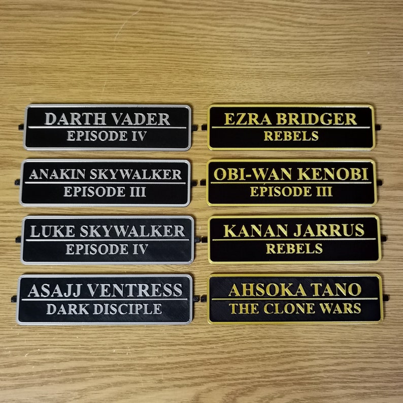 May include: Six black and gold rectangular nameplates with gold lettering. The nameplates feature characters from the Star Wars franchise, including Darth Vader, Ezra Bridger, Anakin Skywalker, Obi-Wan Kenobi, Luke Skywalker, Kanan Jarrus, Asajj Ventress, and Ahsoka Tano.