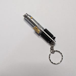 May include: A silver lightsaber keychain with black and gold accents. The keychain has a silver ring attached. The lightsaber has a detailed design, with a button and a textured grip.