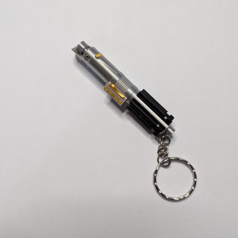 May include: A silver lightsaber keychain with black and gold accents. The keychain has a silver ring attached. The lightsaber has a detailed design, with a button and a textured grip.