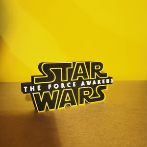 May include: Black and yellow 3D printed Star Wars logo with the text "The Force Awakens" on a yellow background.