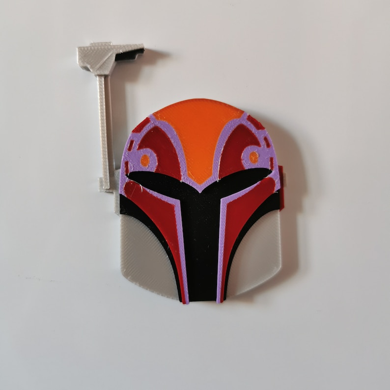 May include: A 3D printed Mandalorian helmet with a red, orange, and purple design. The helmet has a gray base and a small, gray, 3D printed antenna on top.