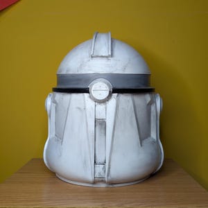 May include: A white and gray helmet with a rounded top and a detailed front design. The helmet has a dark gray band around the middle and is sitting on a wooden surface. The helmet is a replica of a Clone Trooper helmet.