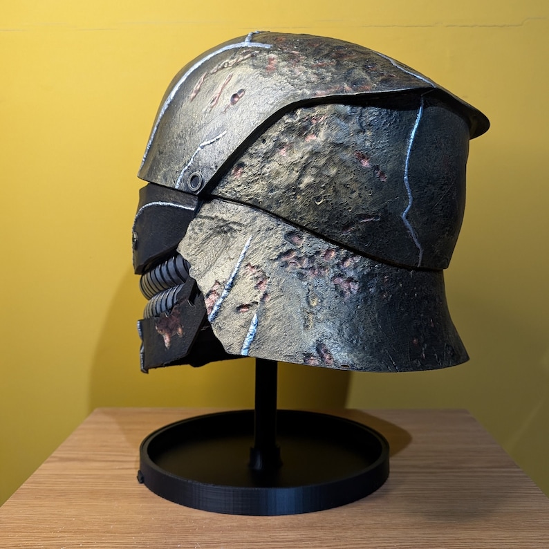 May include: A detailed, weathered, gray and brown helmet with a black face mask and a stand. The helmet has a textured surface and appears to be made of metal.