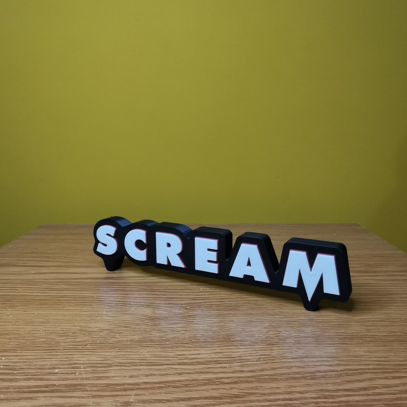 May include: Black and white "SCREAM" sign with red outlining. The sign is made of plastic and has a 3D effect.