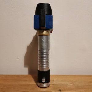 May include: A silver and black lightsaber prop with a blue and gold top. The lightsaber has a ribbed handle and a black grip.