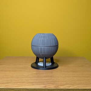 May include: A gray, spherical Death Star-shaped planter with a black base and three legs. The planter is sitting on a wooden surface.