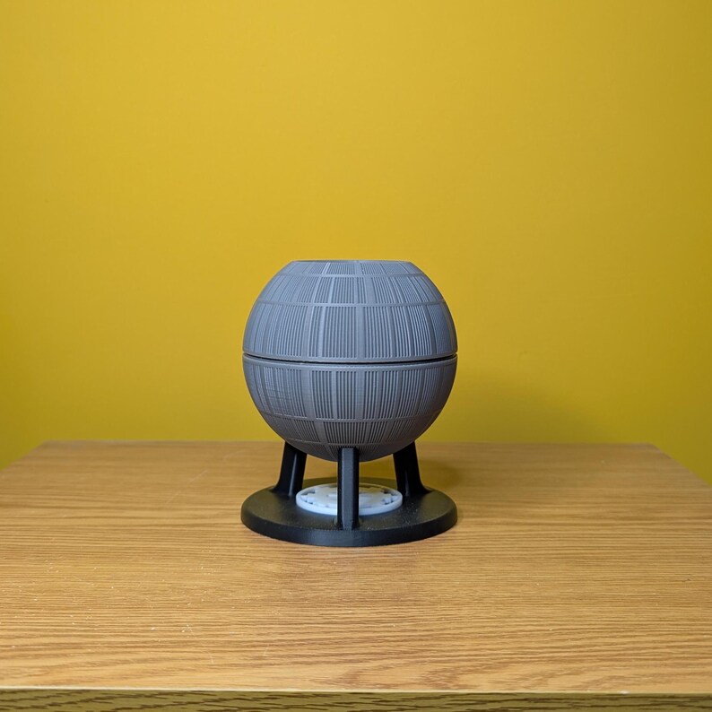 May include: A gray, spherical Death Star-shaped planter with a black base and three legs. The planter is sitting on a wooden surface.