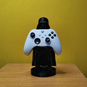 May include: A white Xbox controller is held by a black Darth Vader figurine. The figurine is standing on a black base.
