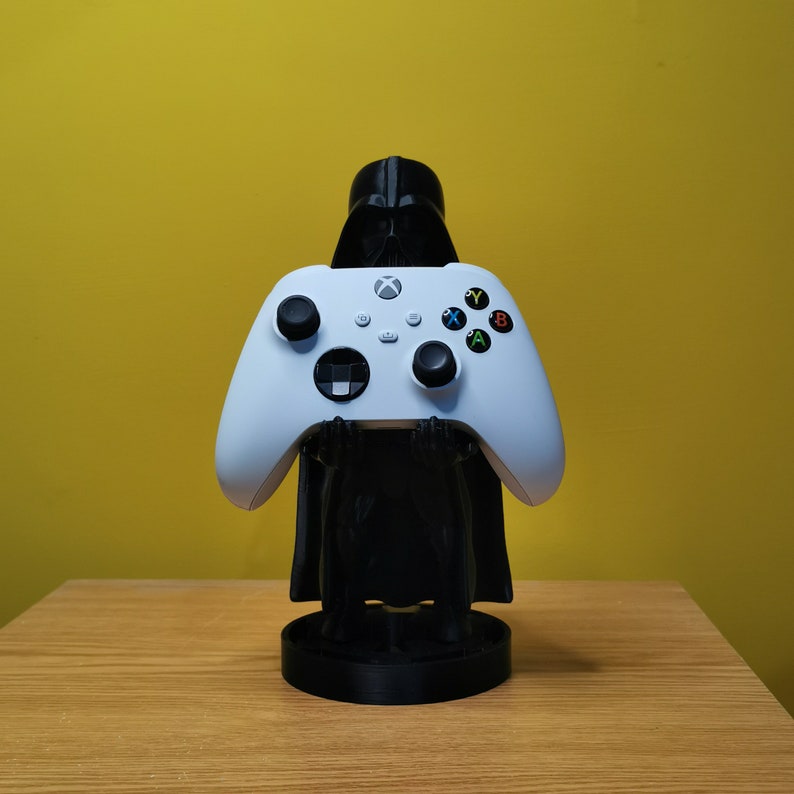 May include: A white Xbox controller is held by a black Darth Vader figurine. The figurine is standing on a black base.