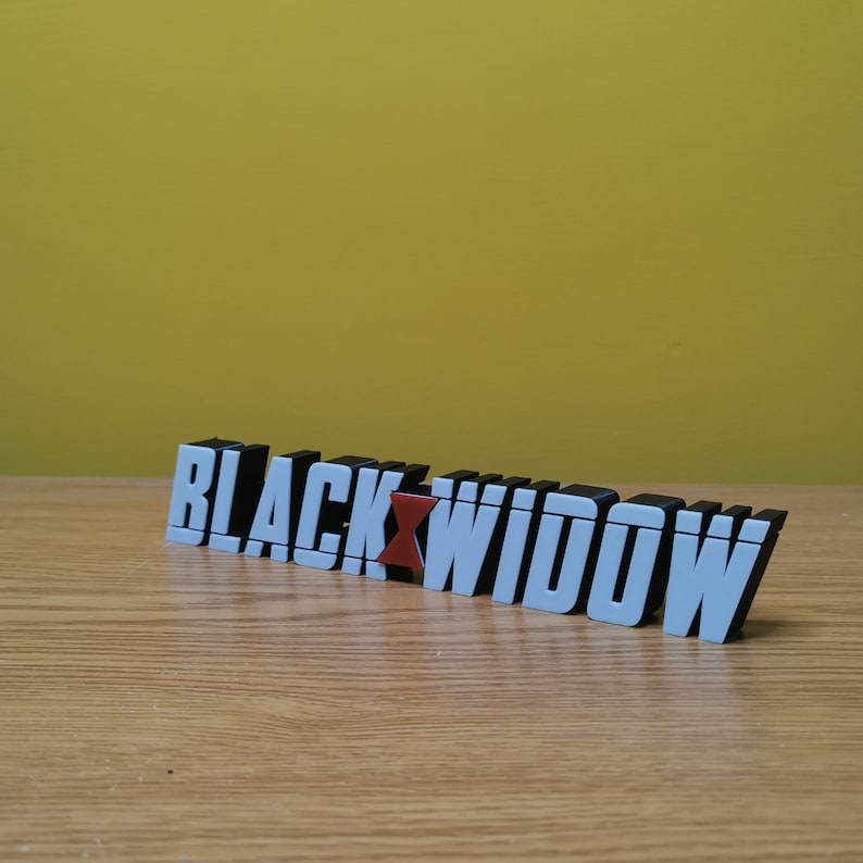 May include: Black Widow logo made of black and white plastic letters with a red widow symbol in the middle. The letters are arranged in a line and are sitting on a wooden surface.