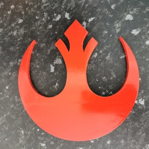 May include: A red Rebel Alliance symbol, also known as the Star Wars Rebellion symbol, is made of a solid material and has a glossy finish.