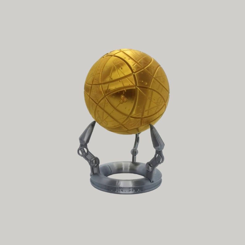 May include: A gold sphere with intricate geometric patterns sits on a silver stand with three legs. The sphere is suspended above the stand.