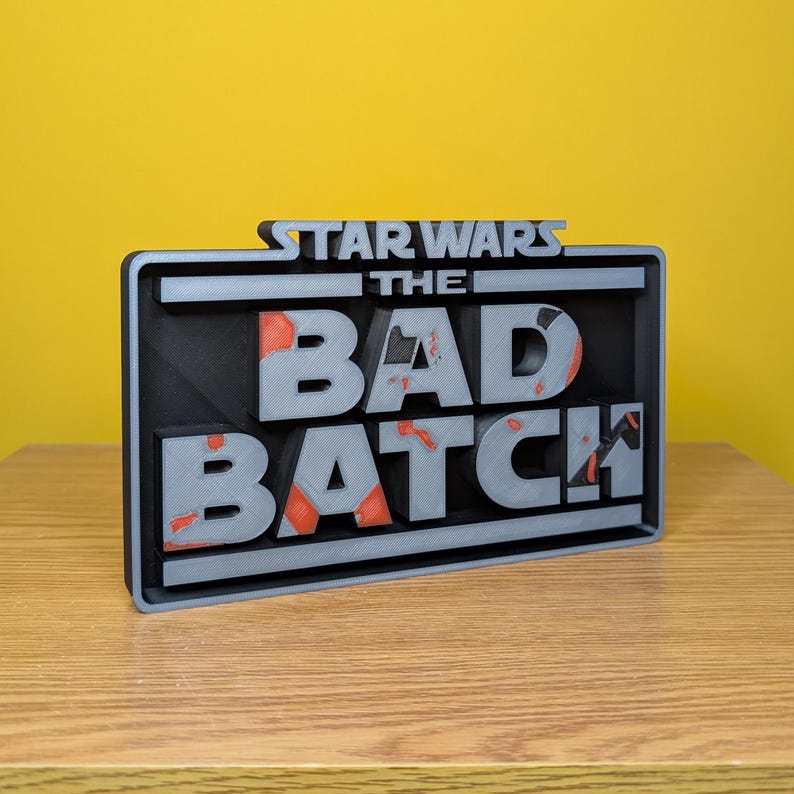 May include: A 3D printed sign with the text "STAR WARS THE BAD BATCH" in a black and gray color scheme. The letters are raised and have a distressed look with red accents.
