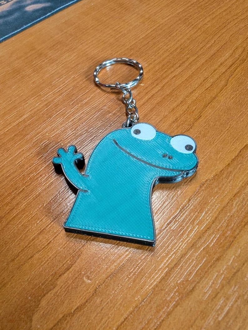 Peut inclure: Un porte-cl&eacute;s bleu sarcelle en forme de grenouille de dessin anim&eacute;. La grenouille a de grands yeux blancs et l&egrave;ve une main. Le porte-cl&eacute;s est attach&eacute; &agrave; un anneau en m&eacute;tal argent&eacute;.
