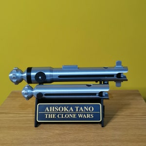 May include: A silver lightsaber prop with a black handle and a silver stand. The stand has a plaque that reads "Ahsoka Tano The Clone Wars".
