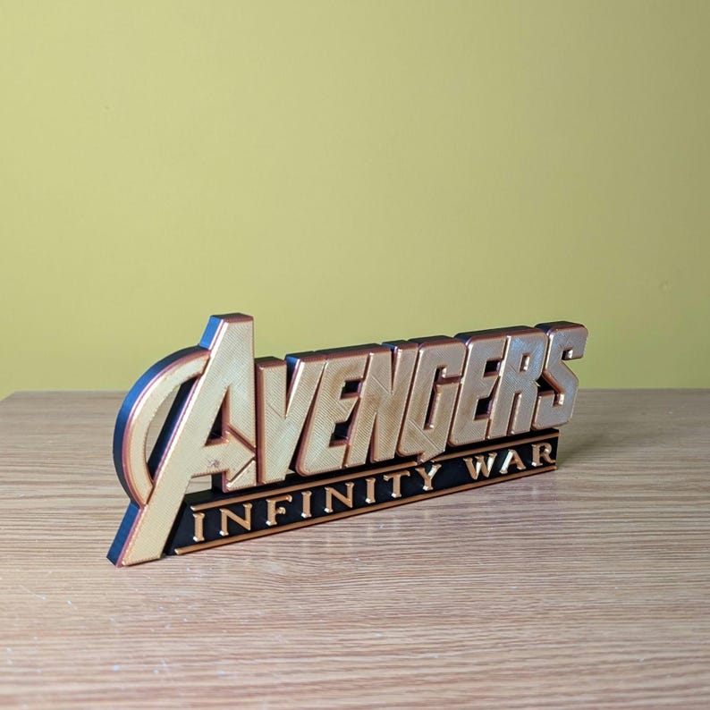 May include: A gold and black Avengers Infinity War logo. The word "Avengers" is in gold, with a black outline, and the words "Infinity War" are in black. The logo is on a wooden surface.