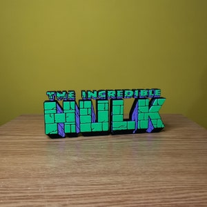 K&ouml;nnte beinhalten: Ein 3D-gedrucktes Schild mit der Aufschrift "THE INCREDIBLE HULK". Die Buchstaben sind gr&uuml;n und lila mit schwarzem Umriss. Das Schild steht auf einer Holzoberfl&auml;che.