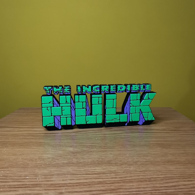 K&ouml;nnte beinhalten: Ein 3D-gedrucktes Schild mit der Aufschrift "THE INCREDIBLE HULK". Die Buchstaben sind gr&uuml;n und lila mit schwarzem Umriss. Das Schild steht auf einer Holzoberfl&auml;che.