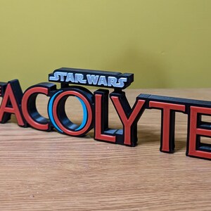 May include: A black and red 3D printed sign that reads "STAR WARS THE ACOLYTE". The sign has a blue and black circle in the middle of the word "ACOLYTE".