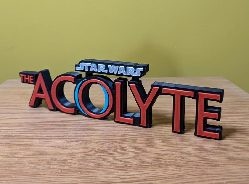 May include: A black and red 3D printed sign that reads "STAR WARS THE ACOLYTE". The sign has a blue and black circle in the middle of the word "ACOLYTE".