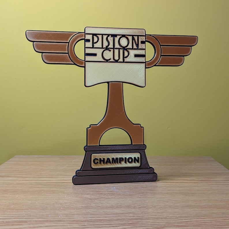 May include: A brown and gold trophy with wings, a piston, and the words "PISTON CUP" on the front. The base of the trophy says "CHAMPION".