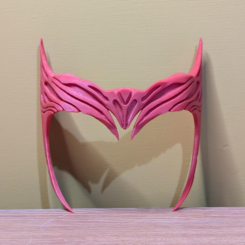 May include: A pink plastic headband with a detailed, intricate design. The headband is shaped like a crown with two points extending down on either side.