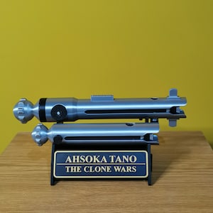 May include: A silver lightsaber prop with a black handle and a silver stand. The stand has a plaque that reads "Ahsoka Tano The Clone Wars".