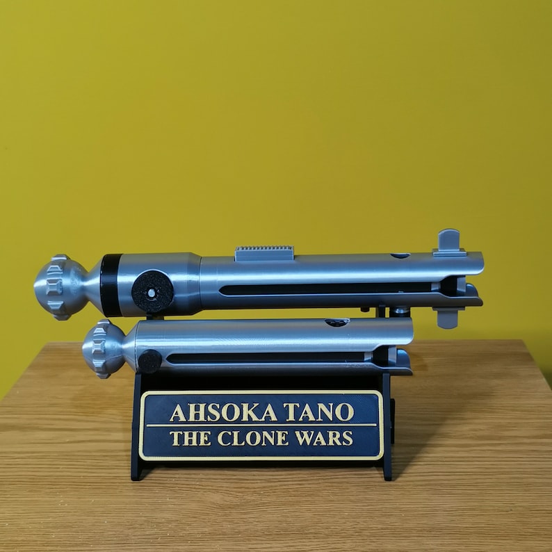 May include: A silver lightsaber prop with a black handle and a silver stand. The stand has a plaque that reads "Ahsoka Tano The Clone Wars".