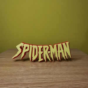 Puede incluir: Un letrero en 3D de color amarillo y rojo que dice "SPIDER-MAN". El letrero está hecho de espuma y tiene un aspecto ligeramente desgastado.