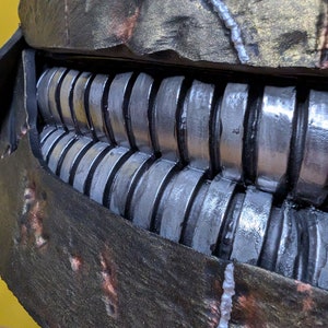 May include: Close-up of a dark gray and brown textured mask with a row of silver metal teeth. The mask has a rough, worn texture with white and copper accents.