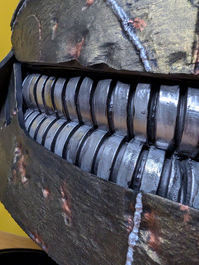 May include: Close-up of a dark gray and brown textured mask with a row of silver metal teeth. The mask has a rough, worn texture with white and copper accents.
