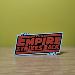 May include: A white, black, and red plastic sign with the text "STAR WARS THE EMPIRE STRIKES BACK".