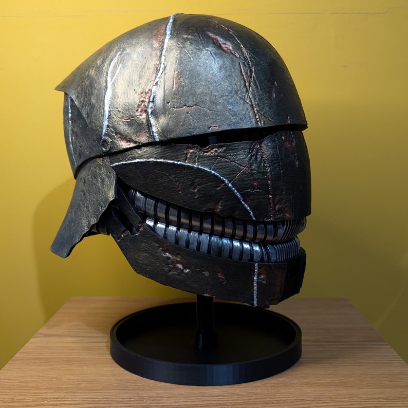 May include: A dark gray and silver helmet with a textured surface and a detailed design. The helmet is on a black stand.