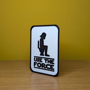 Puede incluir: Un cartel blanco con borde negro y una silueta negra de Darth Vader sentado en un inodoro. El cartel dice "Use the Force" en letras negras.