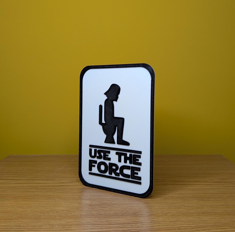 Puede incluir: Un cartel blanco con borde negro y una silueta negra de Darth Vader sentado en un inodoro. El cartel dice "Use the Force" en letras negras.