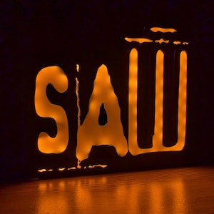 May include: A lit-up sign with the word "SAW" in a stylized font. The letters are outlined in black and the inside is illuminated with a warm orange glow.