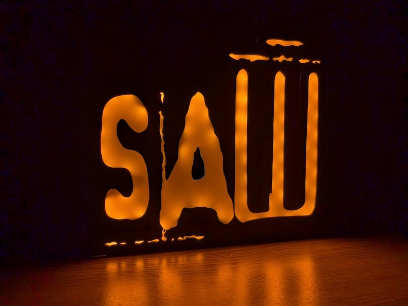 May include: A lit-up sign with the word "SAW" in a stylized font. The letters are outlined in black and the inside is illuminated with a warm orange glow.