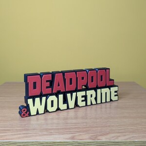 Może przedstawiać: Tr&oacute;jwymiarowy znak z napisem "DEADPOOL & WOLVERINE" w czerwonych i ż&oacute;łtych literach. Litery są ułożone jedna na drugiej, a na g&oacute;rze znajduje się słowo "DEADPOOL".
