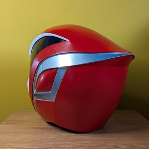 May include: A red and silver plastic helmet with a visor. The helmet is sitting on a wooden surface.