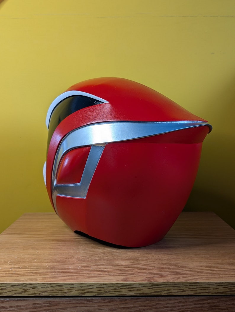 May include: A red and silver plastic helmet with a visor. The helmet is sitting on a wooden surface.