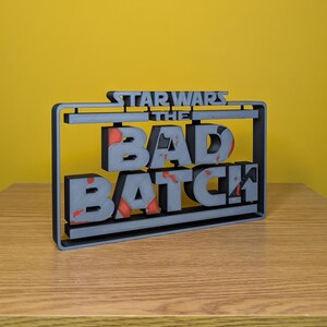 May include: A gray 3D printed sign with the text "STAR WARS THE BAD BATCH" in a black frame. The letters are slightly raised and have a distressed look with red paint splatters.