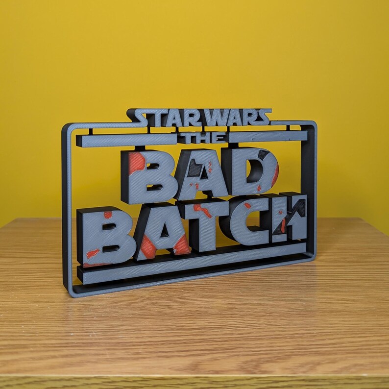 May include: A gray 3D printed sign with the text "STAR WARS THE BAD BATCH" in a black frame. The letters are slightly raised and have a distressed look with red paint splatters.