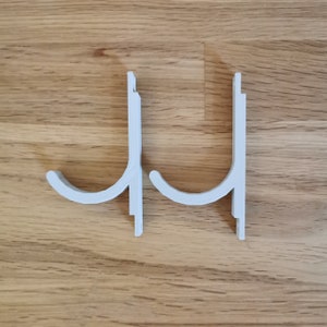 May include: Two white plastic hooks mounted on a wooden surface. The hooks are designed to hang items on a wall.
