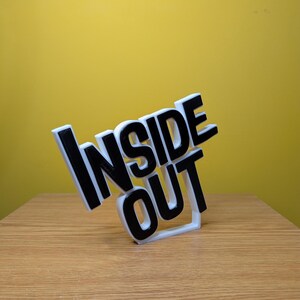 May include: A white 3D printed sign with the words "Inside Out" in black lettering. The sign is tilted to the side and is sitting on a wooden surface.
