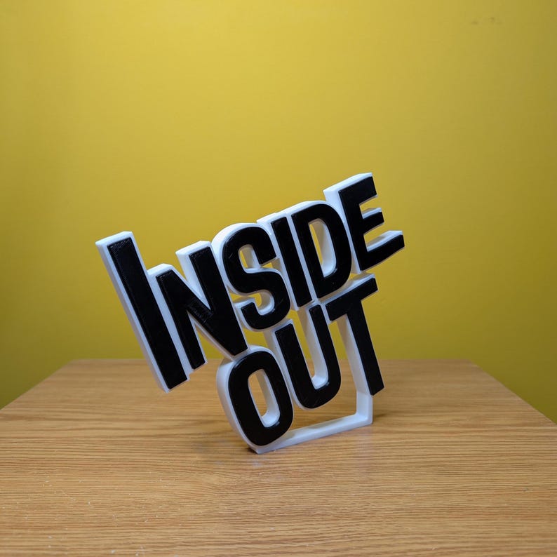 May include: A white 3D printed sign with the words "Inside Out" in black lettering. The sign is tilted to the side and is sitting on a wooden surface.