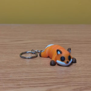 May include: Orange and white fox keychain with a silver key ring. The fox is lying on its side with its head resting on its paws.