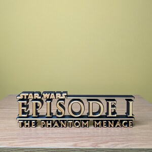 May include: A black and gold 3D printed Star Wars Episode I: The Phantom Menace logo. The logo is on a wooden surface.