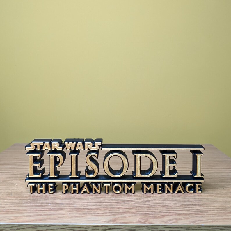 May include: A black and gold 3D printed Star Wars Episode I: The Phantom Menace logo. The logo is on a wooden surface.