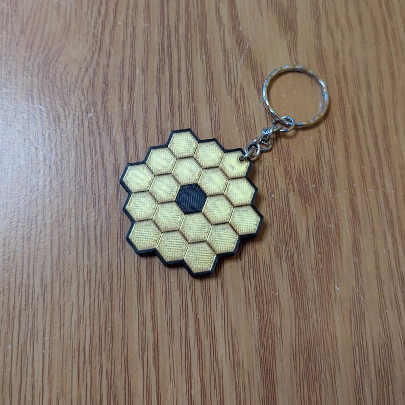 May include: A keychain featuring a gold and black geometric design, resembling a honeycomb pattern. The central portion is a dark circle, surrounded by hexagonal shapes. The keychain has a silver-colored ring.